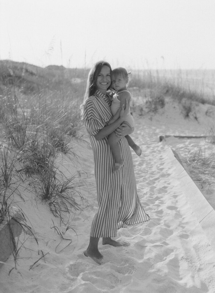 A mother in a striped tunic holds her little baby in a swimsuit on the beach in Destin, Florida. 