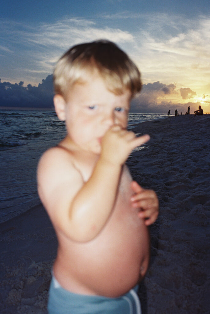 A little boy in blue swim trunks pouting with his thumb in his mouth while the sun sets on the beach in Panama City Beach, Florida.