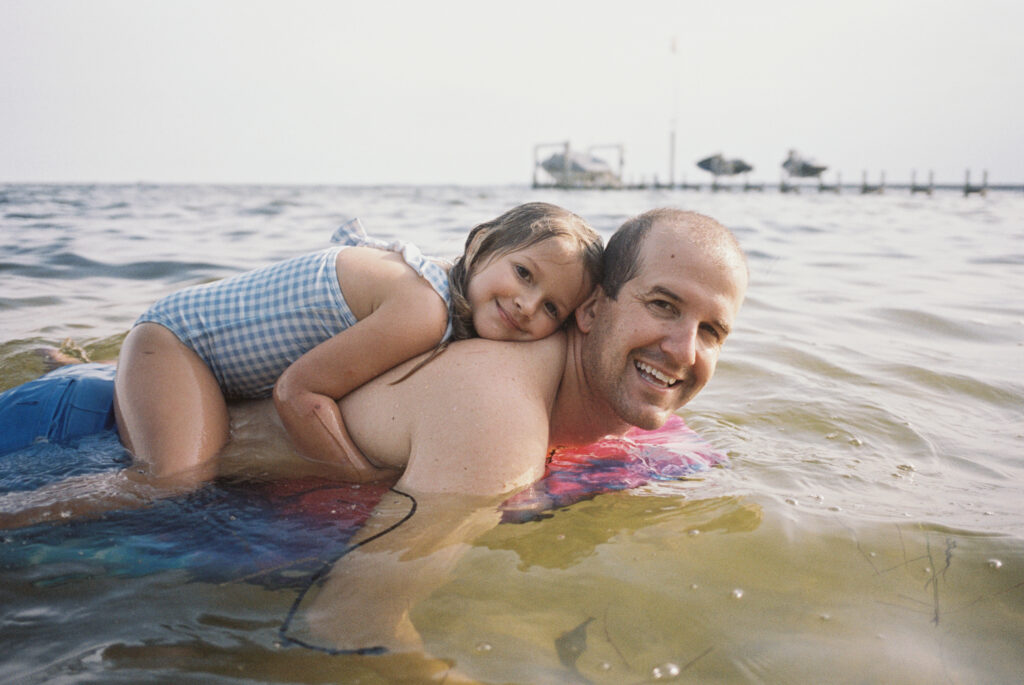 A dad swimming with his daughter floating on his back in her blue gingham swimsuit at Blackpoint in Shalimar, Florida.