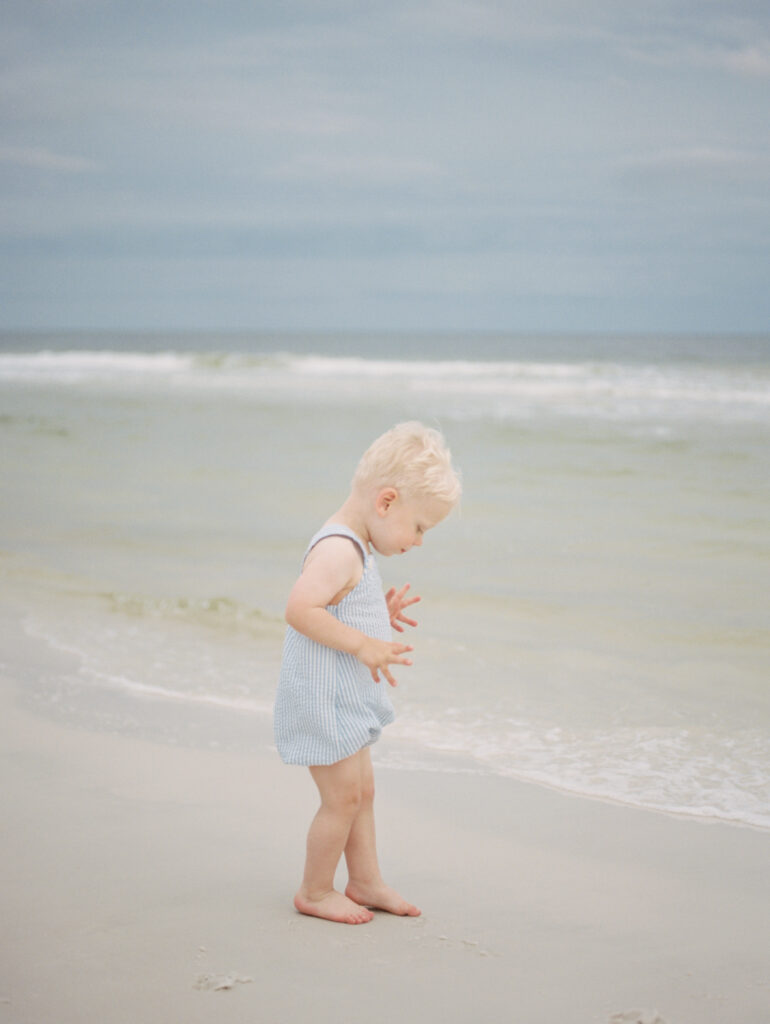 A blonde hair boy in a blue and white overalls touches his toes in the water on the beach in Watercolor, Florida.