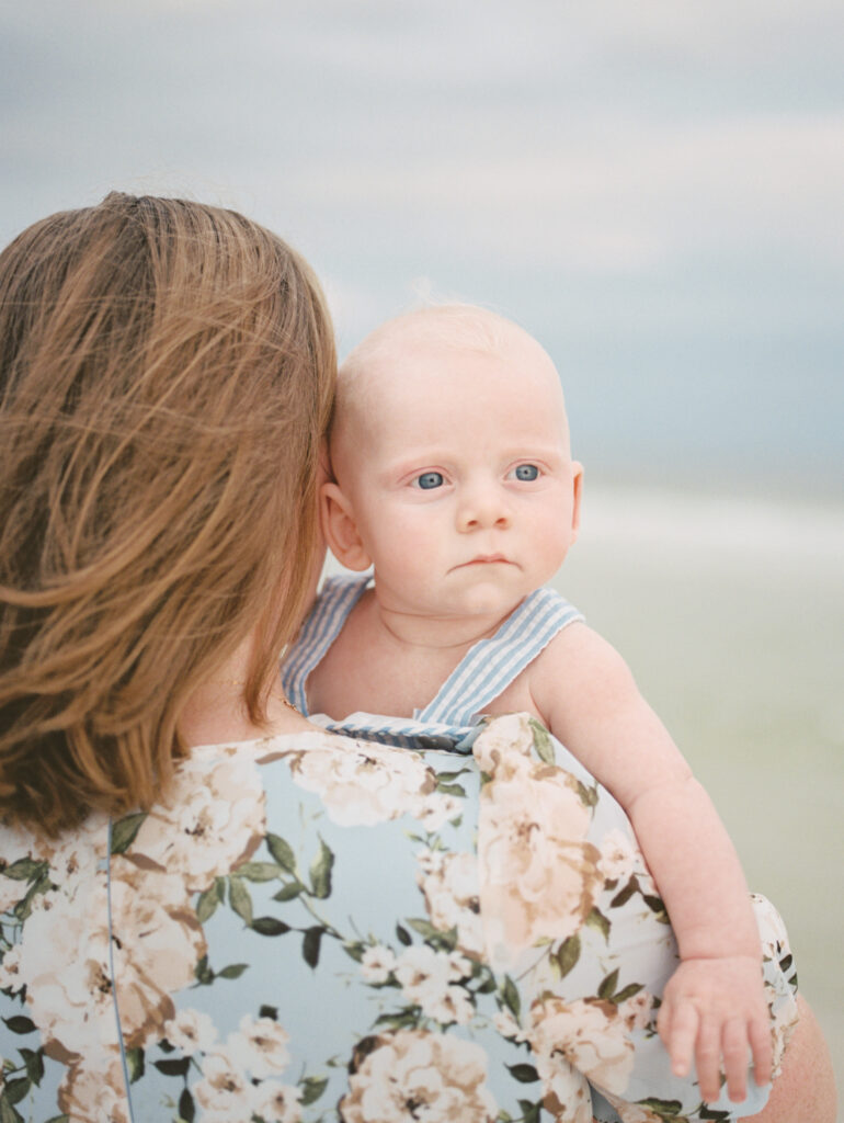 A mother in a floral dress looks out onto the sea while holding her blue eyed baby boy in his blue and white striped pinafore.