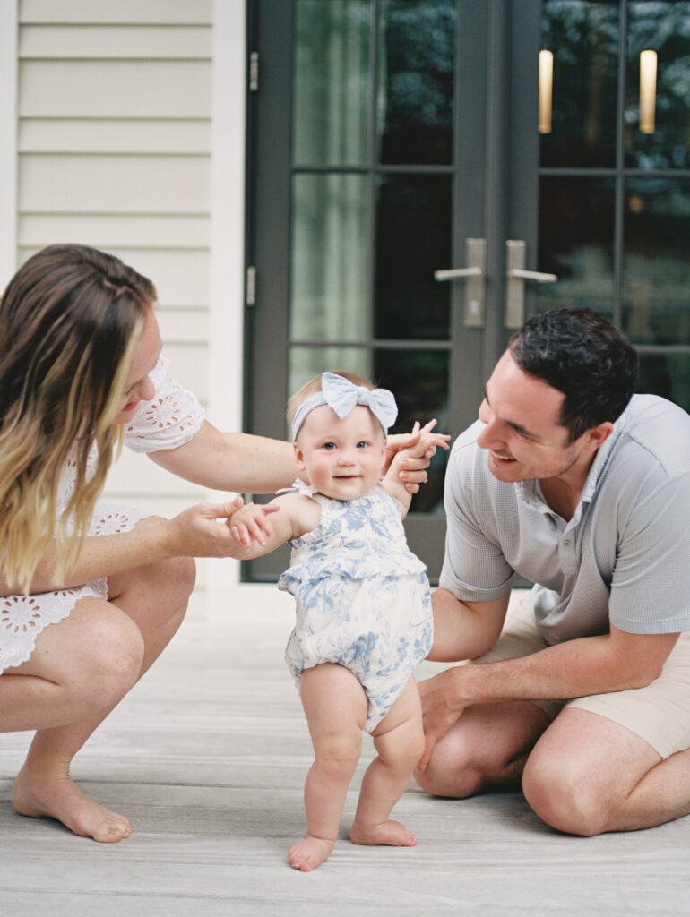 A little baby girl in a blue and white romper with her parents on the porch of their beach house in Watercolor, Florida.