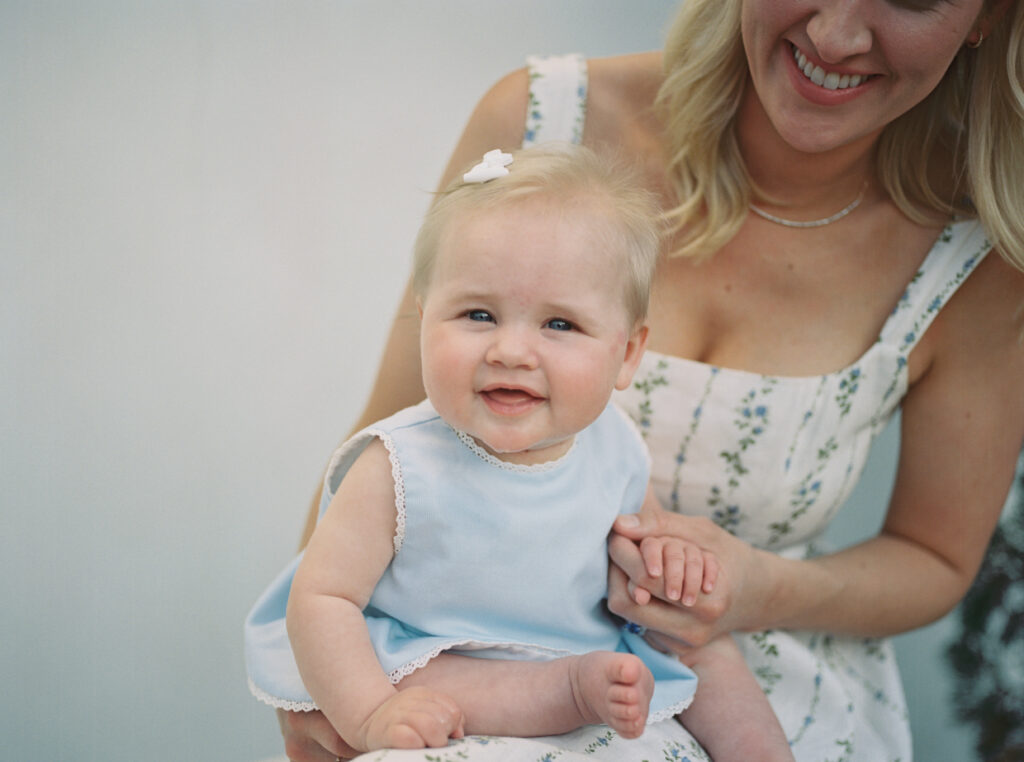 A blonde hair blue eyed baby in a blue dress sits in her moms lap outside their Alys Beach, Florida home.