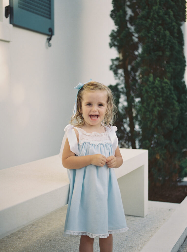 A little girl in a blue and white heirloom dress stands smiling at her parents while on vacation in Alys Beach, Florida.