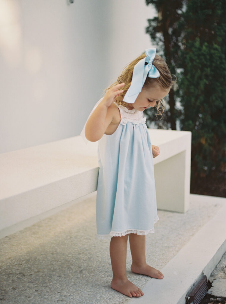 A little girl with a large blue bow in her hair and a blue and white heirloom dress on laughs down at her toes.
