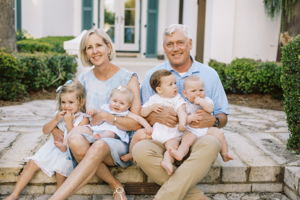 Grandparents sit on the stairs in front of their Alys Beach, Florida home while holding all of their grand babies.