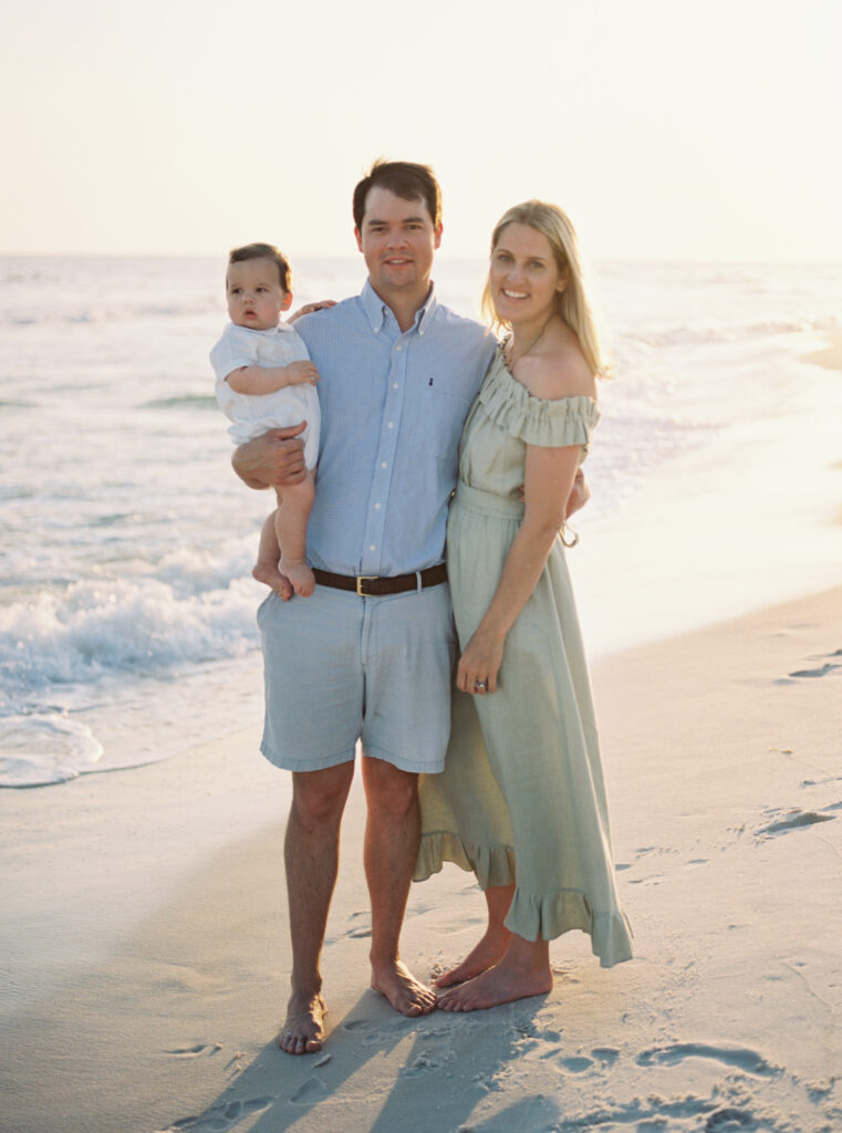 A mother with a green midi dress on and a father with a blue polo shirt on holds their young son at sunset on the beach in 30a.