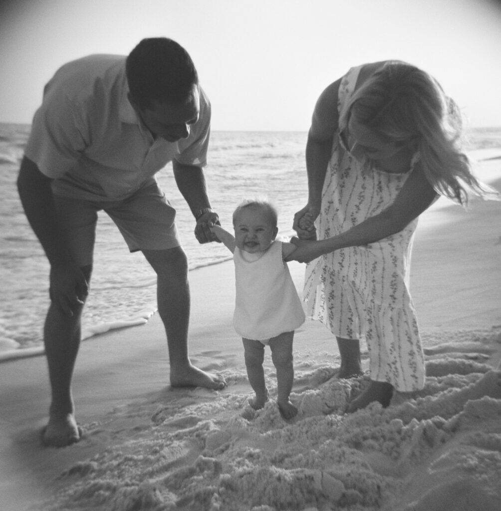 A little girl laughs with her tongue out as her parents hold her hands on the beach in 30A Florida.