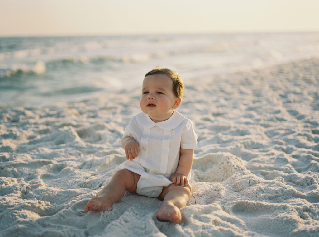 A brown hair little boy in a white heirloom outfit sits on the beach as the sun goes down in Alys Beach, Florida.
