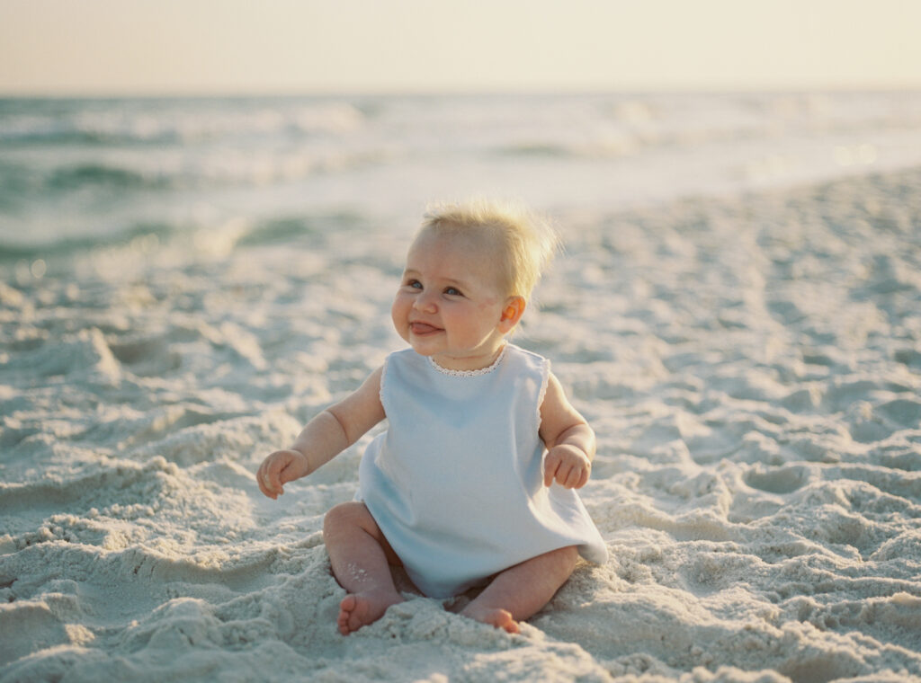 A newborn baby girl sits on the beach smiling at her parents as the sun sets in 30A Florida.
