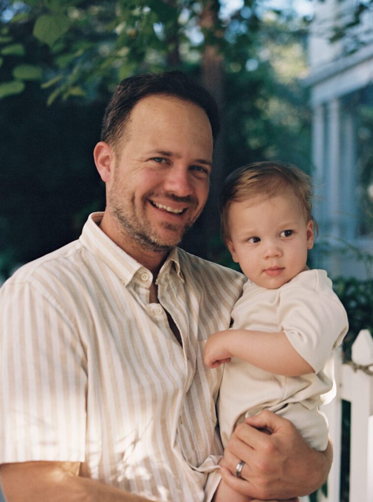 A father in a tan and white striped shirt holds his son who is wearing a natural outfit in Seaside, Florida.