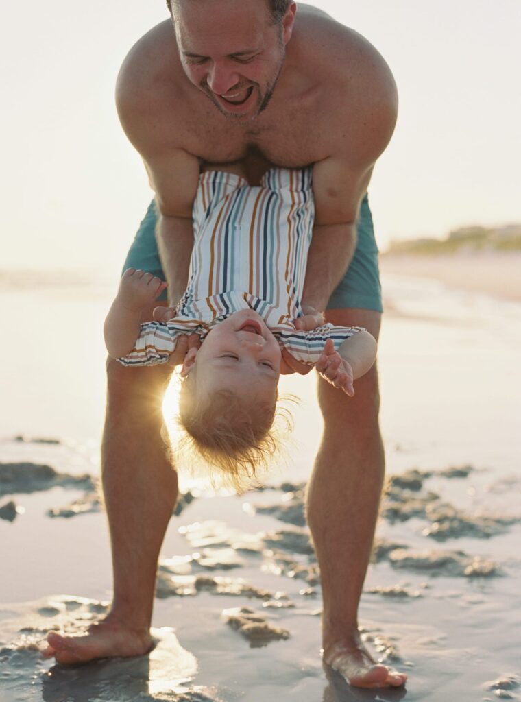 A father bends over and his young son giggles with joy on the beach in 30A Florida.