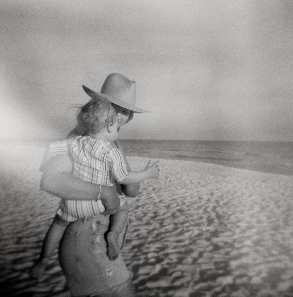 A mother in a natural sunhat walks on the beach in 30A with her newborn son on her hip.