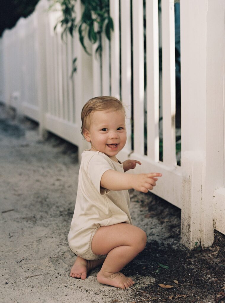 A baby boy in an oat colored oneside tries to steady himself to stand while holding onto a fence in Seaside, Florida.