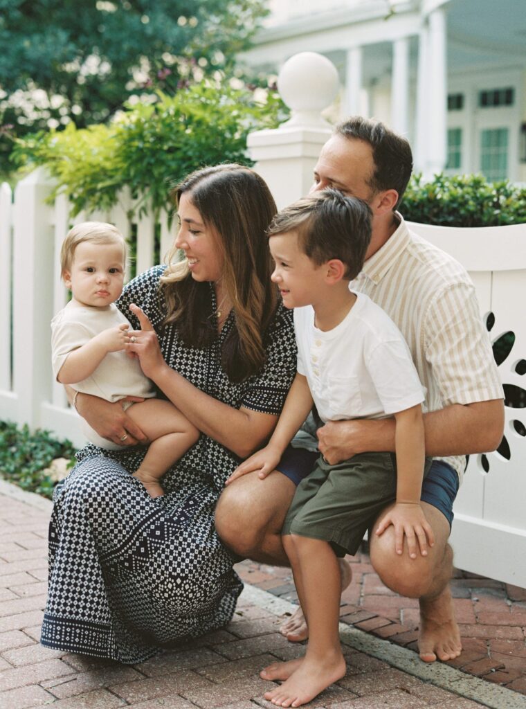 A mother and father crouch down on a sidewalk in Seaside, Florida to hold and laugh with their two young sons.