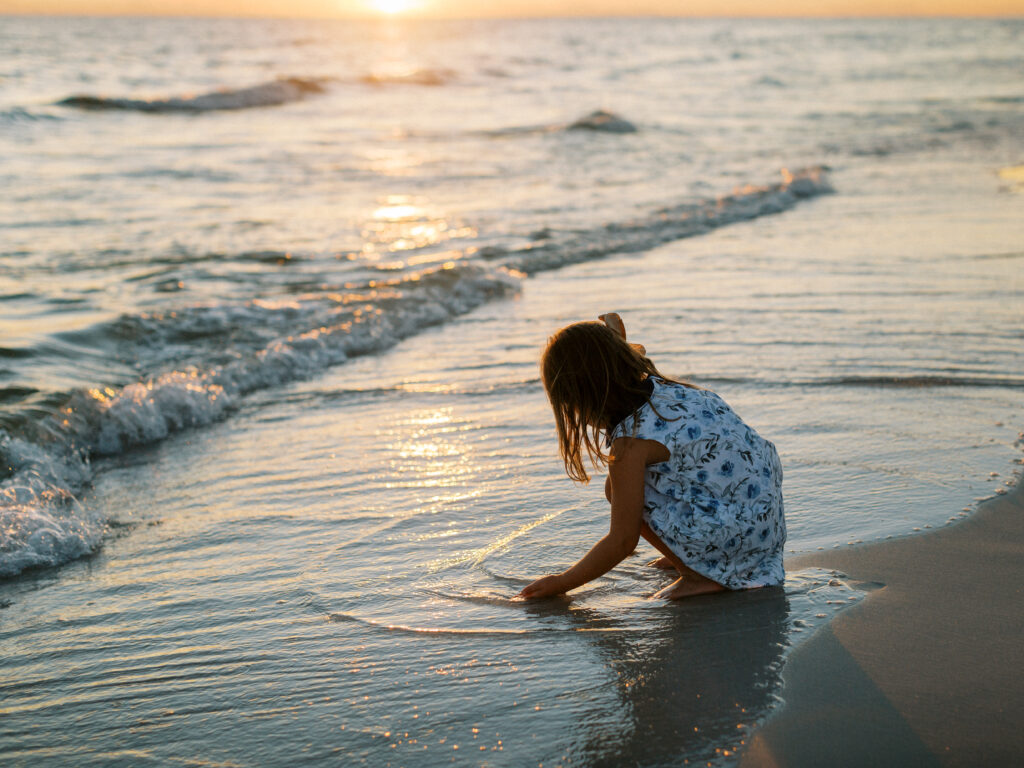 A little girls bends down at the shoreline as the sun sets behind her picking up shells in grayton beach, florida.