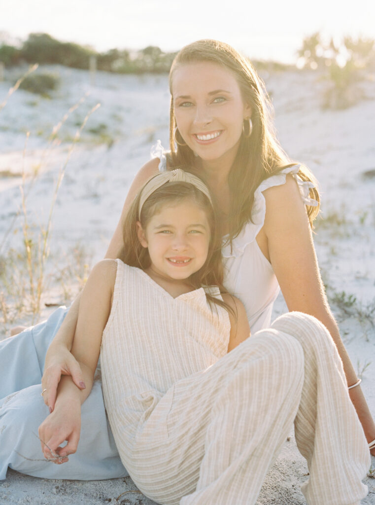 A mother and daughter sitting on the sand with the Grayton Beach State park dunes in the background.