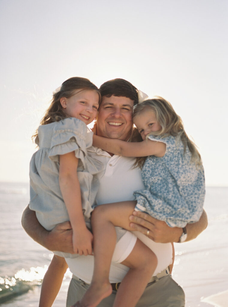 A dad holds his two daughters in his arms while smiling together on the beach in Destin, Florida. 