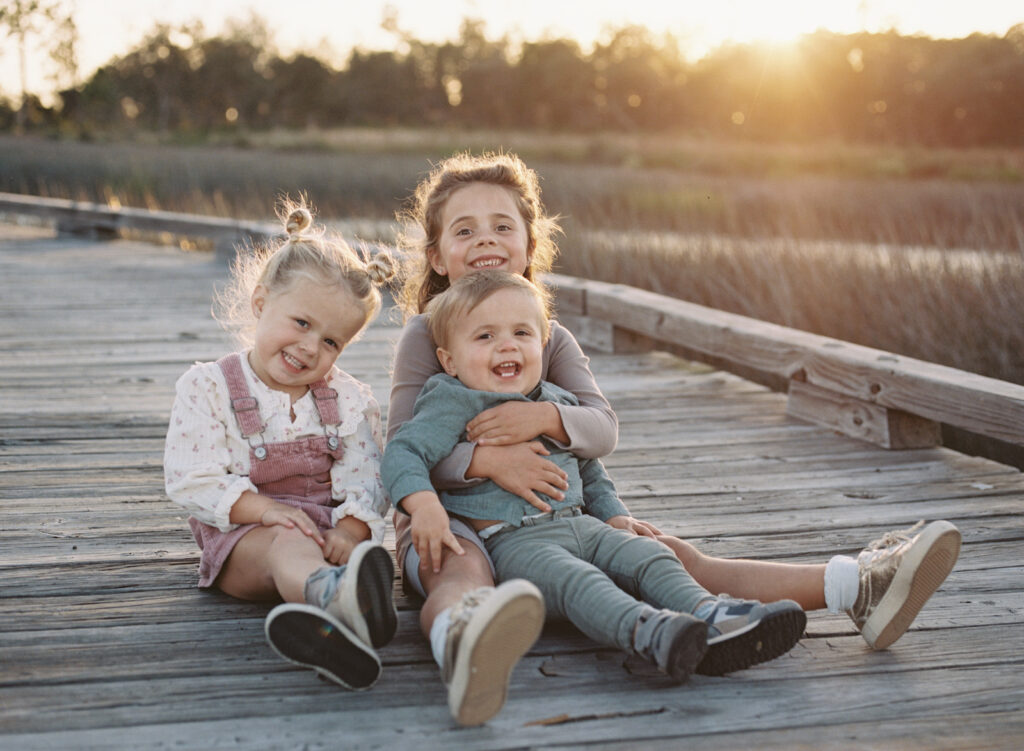 Two sisters and their young brother sitting on a dock on the bay as the sun sets behind them.