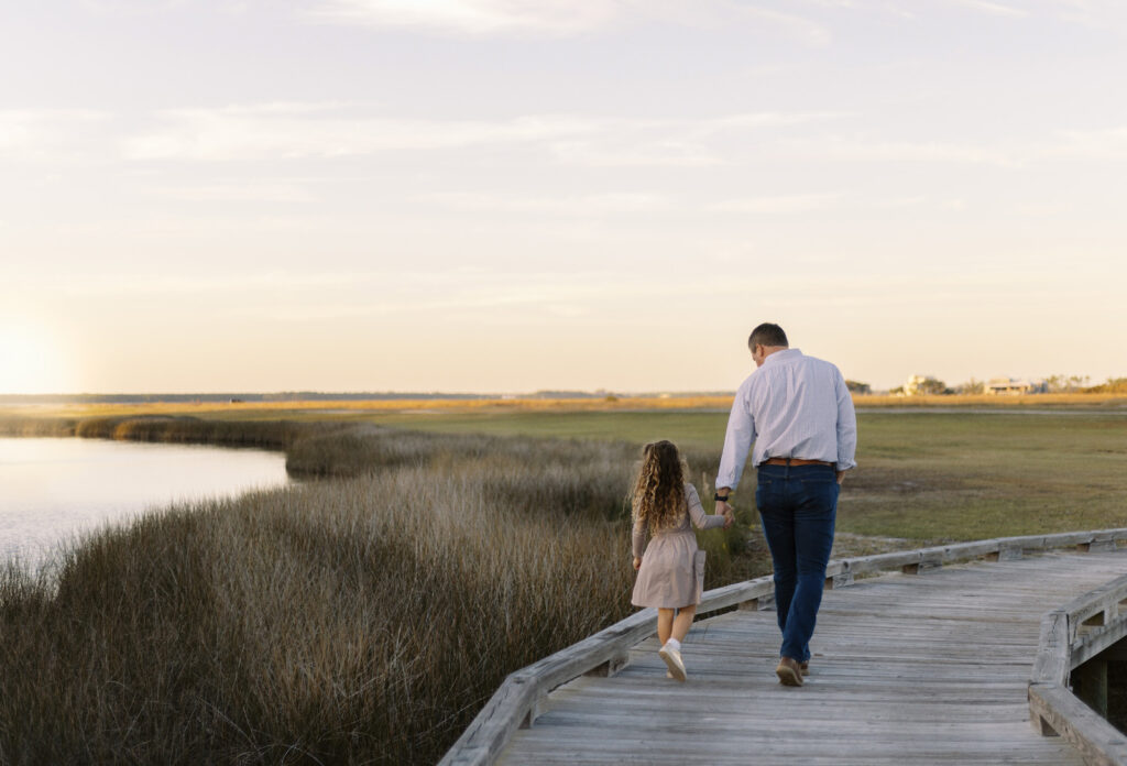 A father and his daughter walking on a dock looking at the bay to their left.