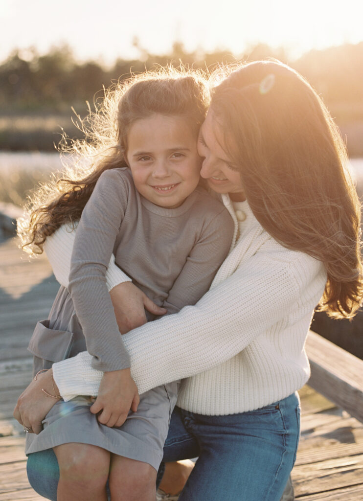 A mother with long brunette hair embracing her daughter that is sitting on her lap while the golden sun shines behind their faces. 
