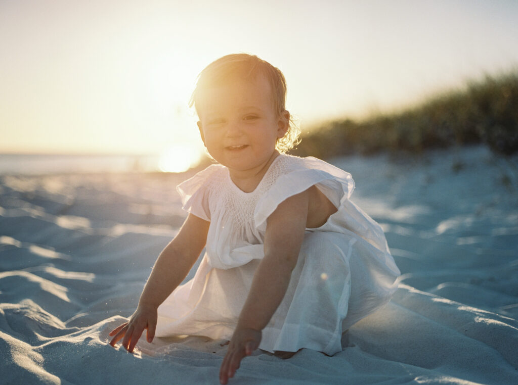 A little girl plays in the sand in a white heirloom dress as the sun sets behind her in Grayton Beach, Florida.