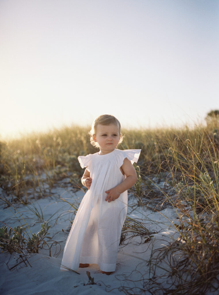 A little girl stands in a white heirloom dress in the sand dunes as the sun sets in Grayton Beach, Florida.