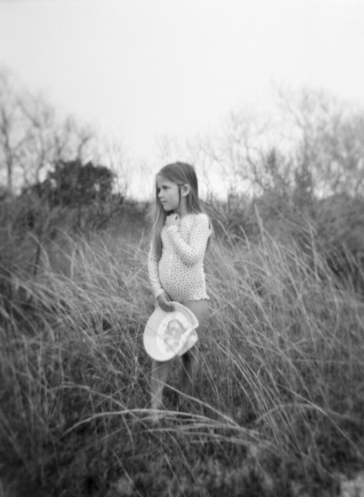 A little girl in a polka dot swimsuit with a beach hat stands among the sea oats in Destin, Florida. 